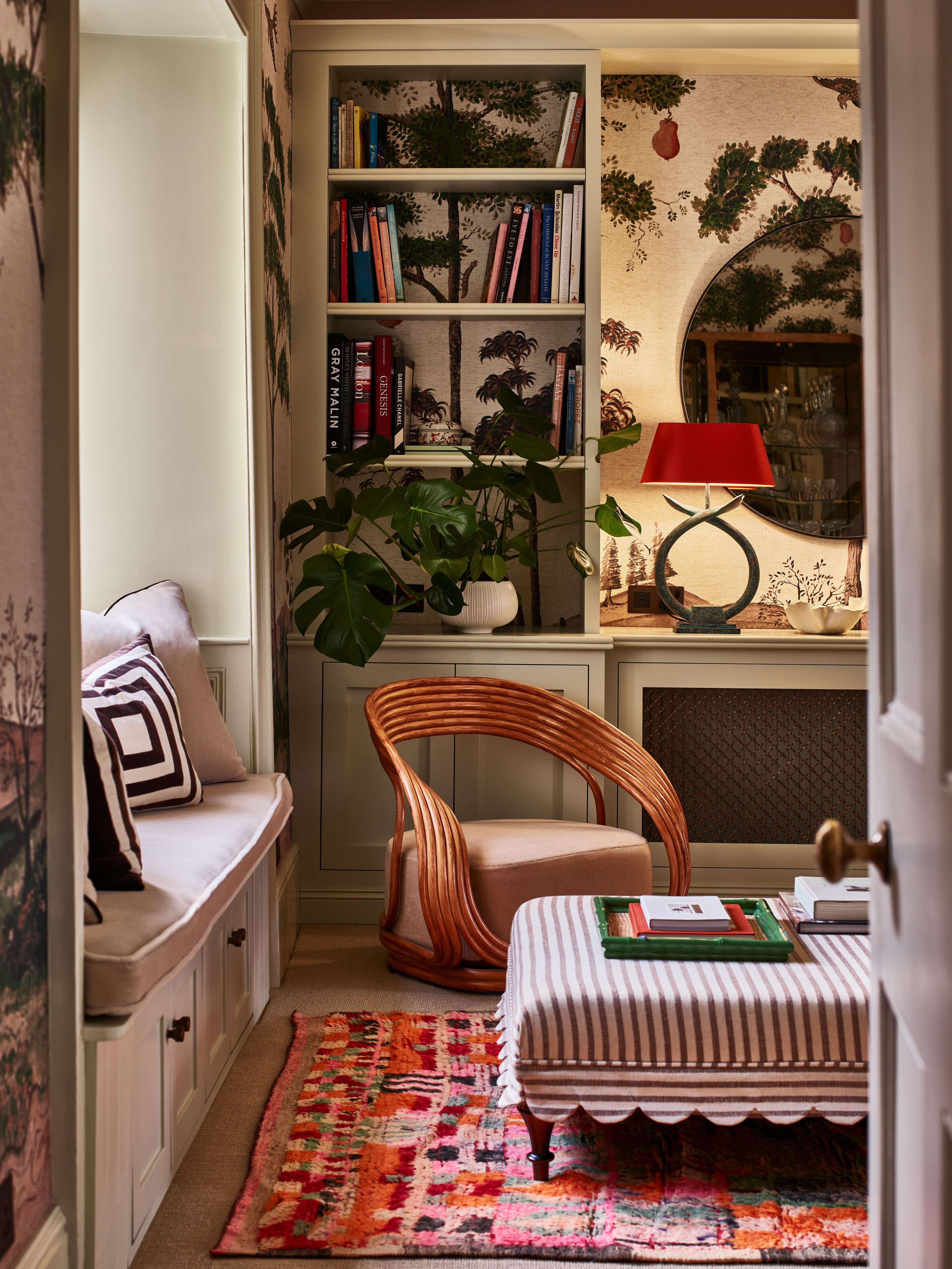 cozy snug room with mural wallpaper and neutral bookshelves styled with a red table lamp, window seat nook with a striped ottoman with scalloped trim