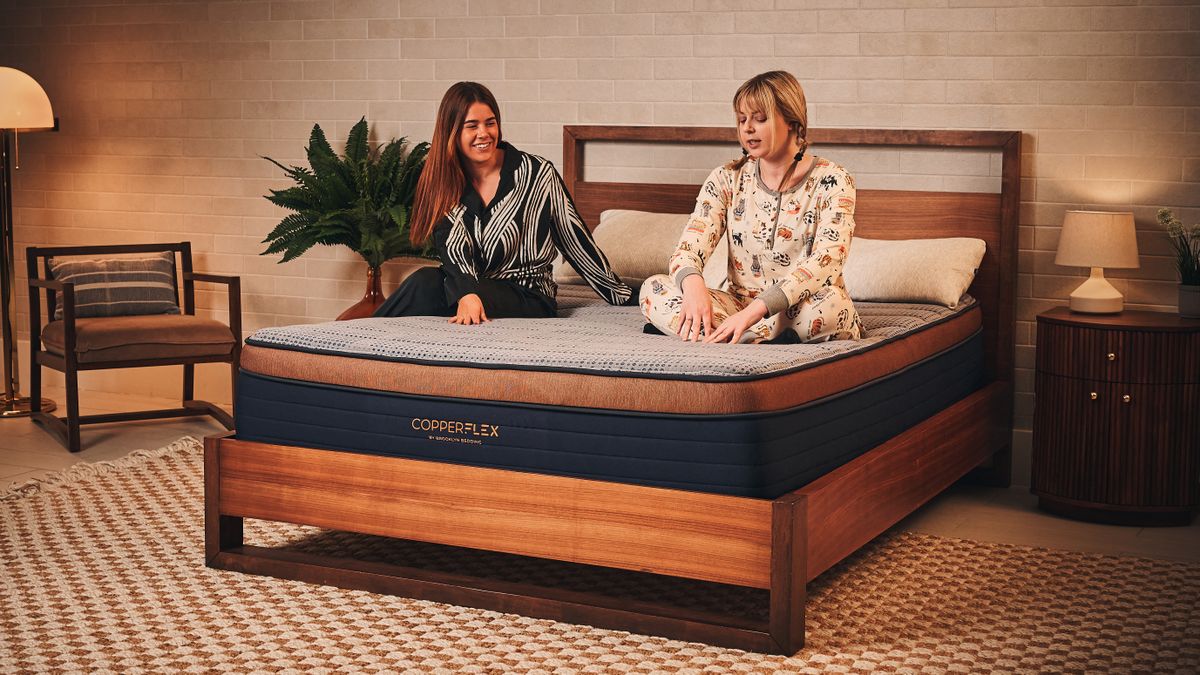 Two women in pyjamas sit on a Brooklyn Bedding CopperFlex mattress. One woman in a black and white patterned shirt sits near the edge, smiling at her friend, while the other woman in patterned pajamas sits cross-legged near the pillows, looking down and talking. The mattress has a textured blue-grey top quilt, a copper-toned border, and a dark blue base displaying the Brooklyn Bedding Copperflex logo. The bed is in a warm, modern bedroom with a checkered rug and a wooden nightstand.