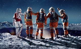 four people in space suits put their arms on one another's shoulders on the surface of the moon while looking down into a large crater