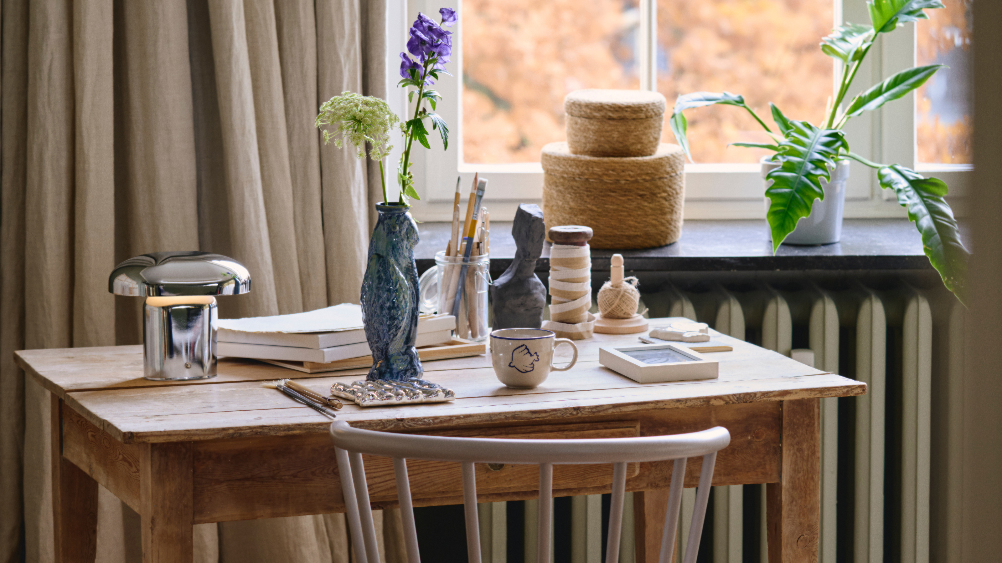 S&oslash;strene Grene chome lamp, fish vase, blue mug, woven storage baskets and weaving materials on a wood table.