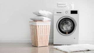 A running washing machine next to a laundry hamper with towels and pillows