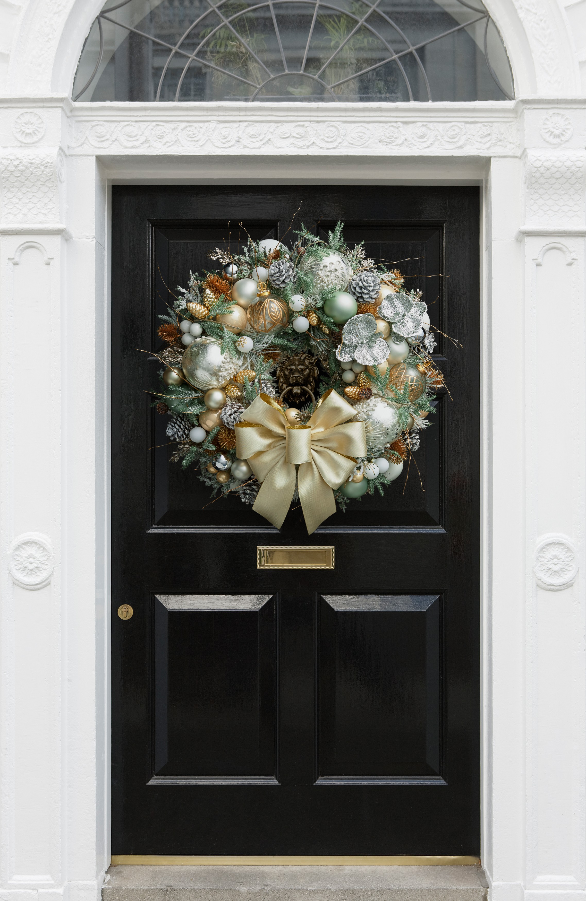 Image of a luxury Christmas wreath on a black front door. The wreath is large with an icy theme. There are mint and champagne colored baubles, foliage, and ribbon.