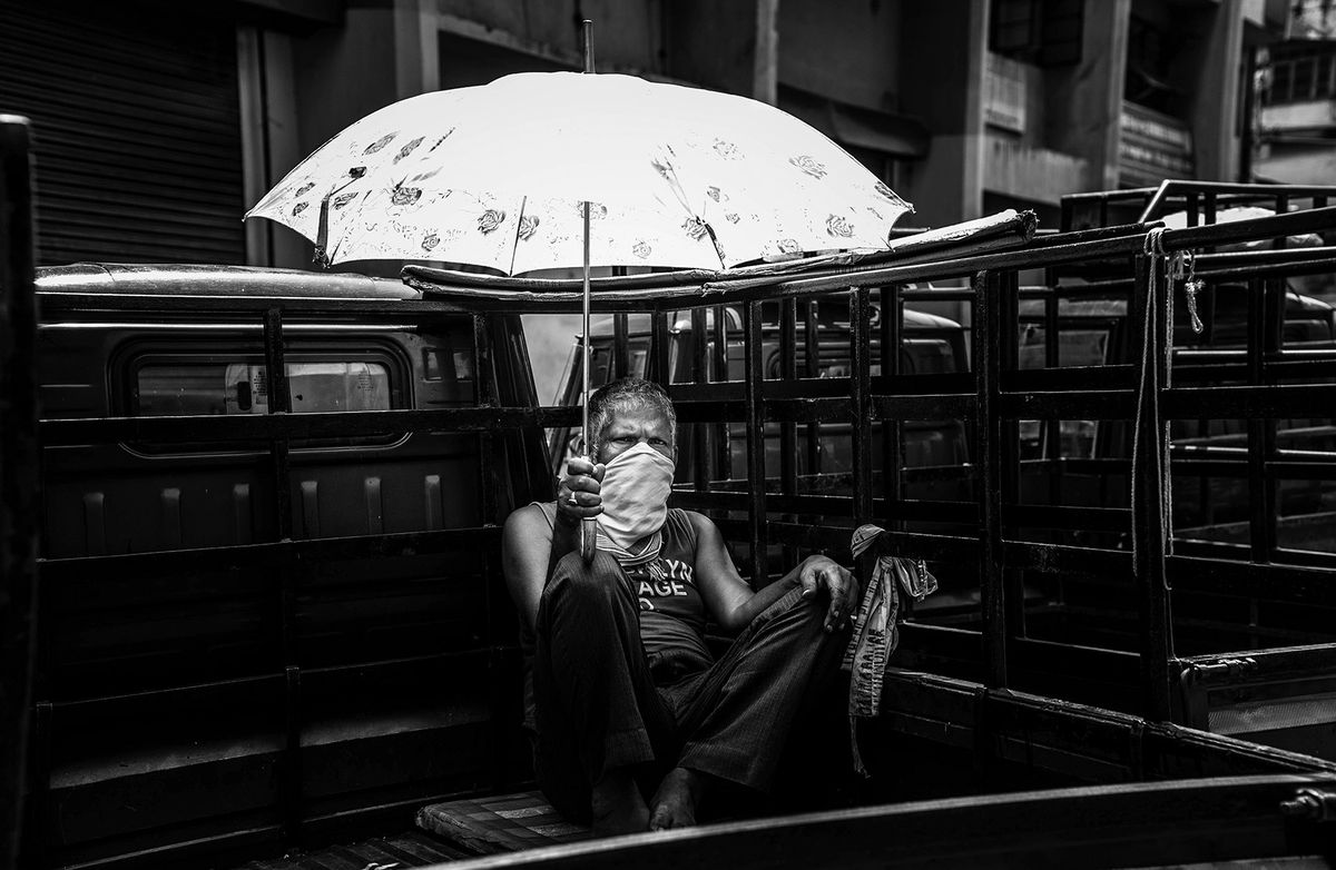 Black-and-white image of a man holding an umbrella while sitting in the back of a van, wearing a COVID mask.