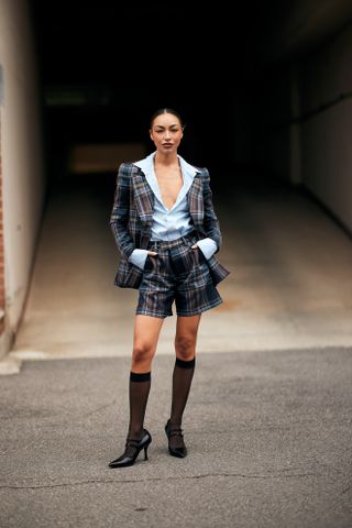 Woman at Paris Fashion Week wearing sheer knee-high socks, black heels, a blue and red plaid blazer with matching shorts and a blue shirt.