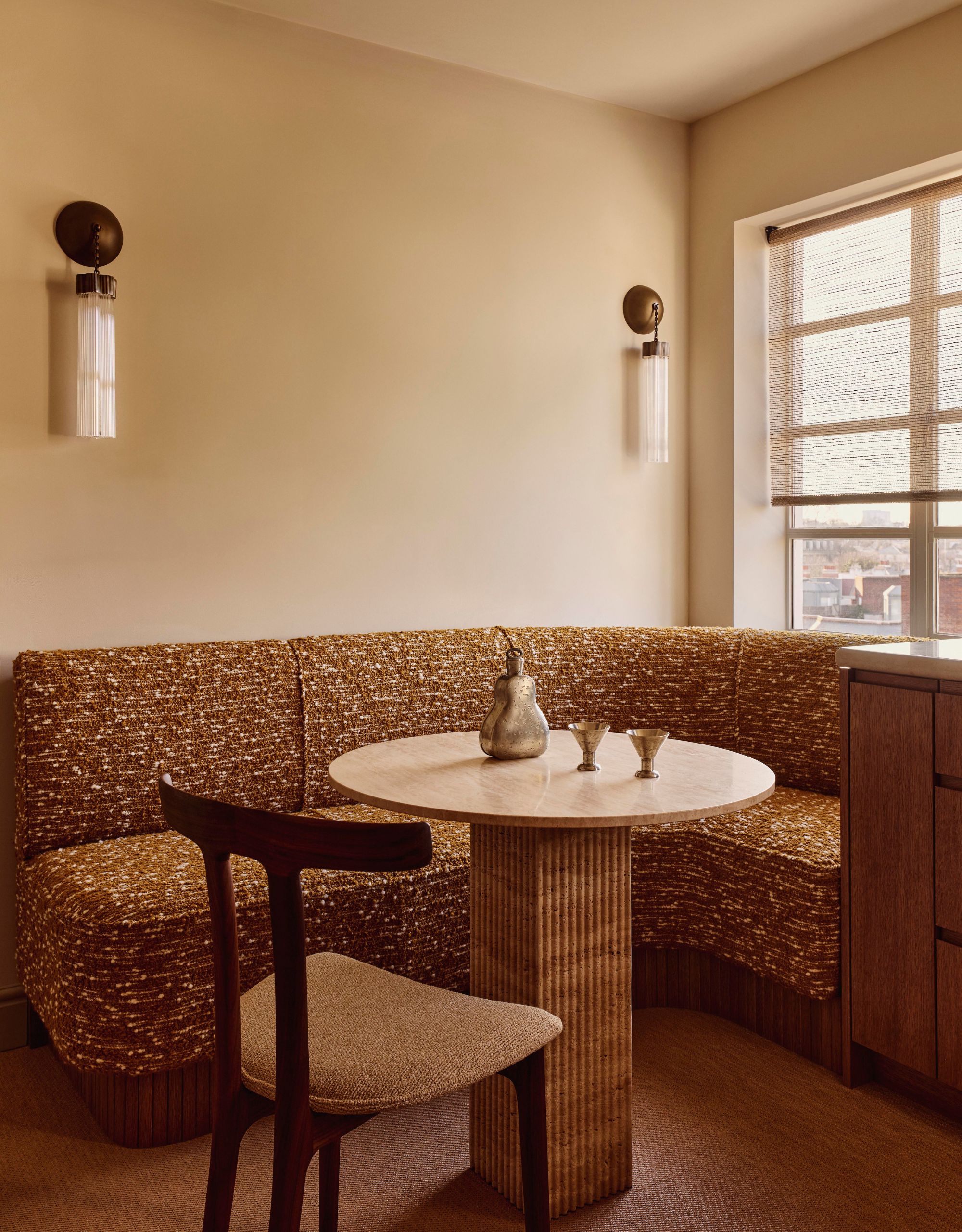A banquette seating corner with ochre textured seating in front of a pedestal dining table with a dining chair on the other side and two wall lights on the neutral wall behind