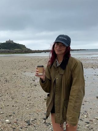 Dua Lipa wearing a green barn jacket with a baseball cap on the beach.