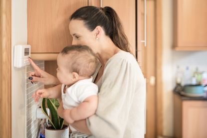 Young Mother holds her baby boy on her arms and shows him how to adjusts the temperature of the household on a thermostat in the kitchen