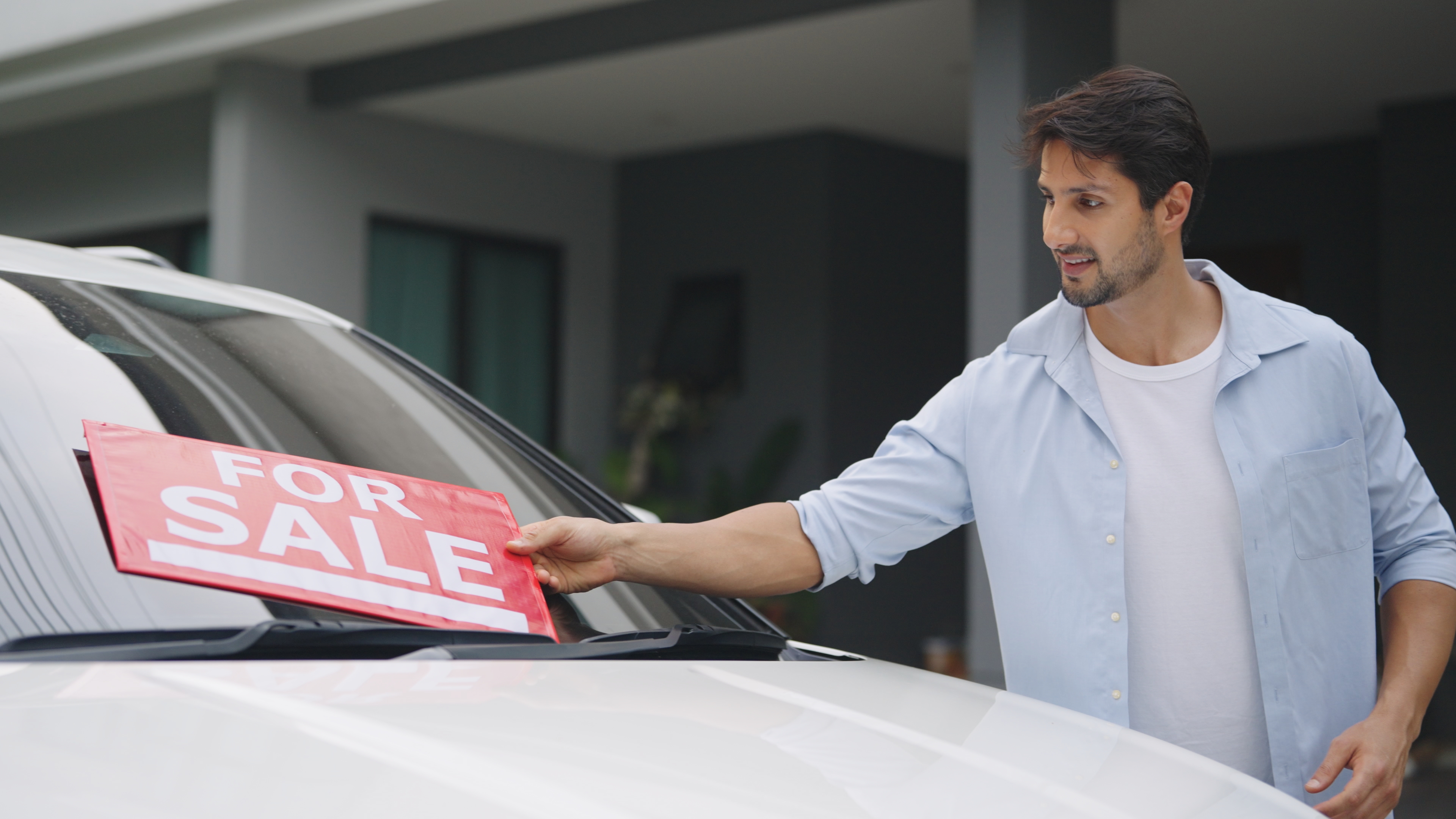 A young man placing a red "For Sale" sign on the windshield in front of a car 