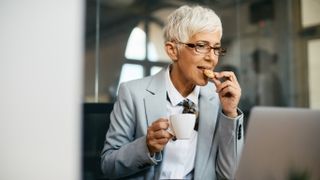 A woman sitting at a table, looking at a laptop screen, biting on a cookie, and holding a cup