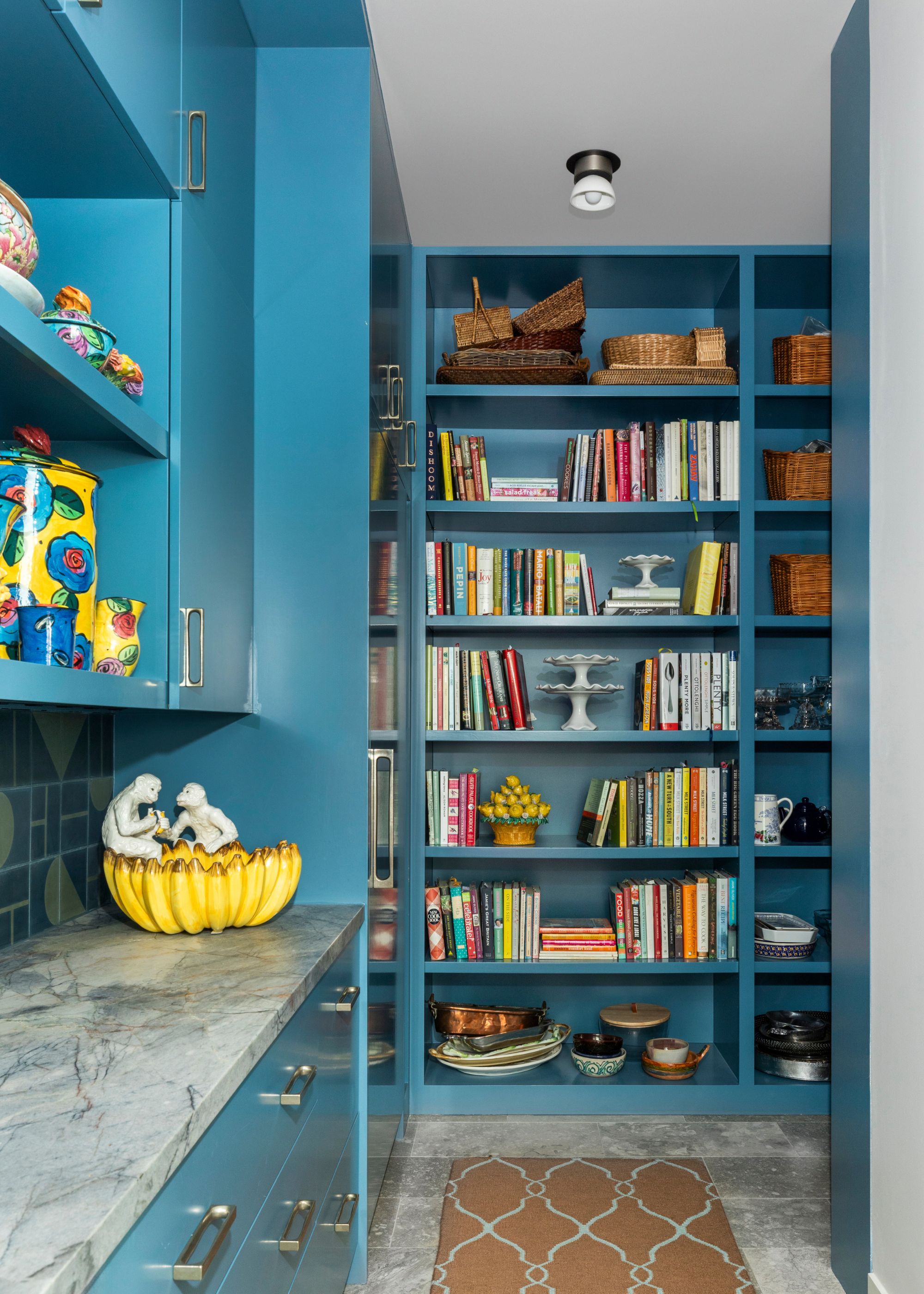 A pantry painted bright blue with built-in shelving, stone countertops and flooring, and colorful decor on the shelves.