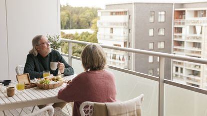 An older couple sit on the balcony of their high-rise and enjoy a meal.