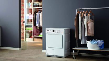 the best tumble dryer, the Miele TDA140 C Freestanding Condenser Tumble Dryer, sat in a wardrobe room next to a rack of clothes, and a cupboard full of clothes and towels beyond, through a glass door