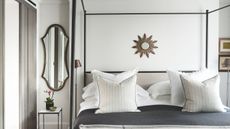 White bedroom with a black four-poster bed, grey bedding with white cushions, and a small circular mirror above the bed. To the left is an oval shaped mirror.