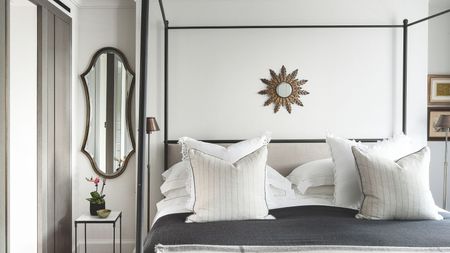 White bedroom with a black four-poster bed, grey bedding with white cushions, and a small circular mirror above the bed. To the left is an oval shaped mirror.