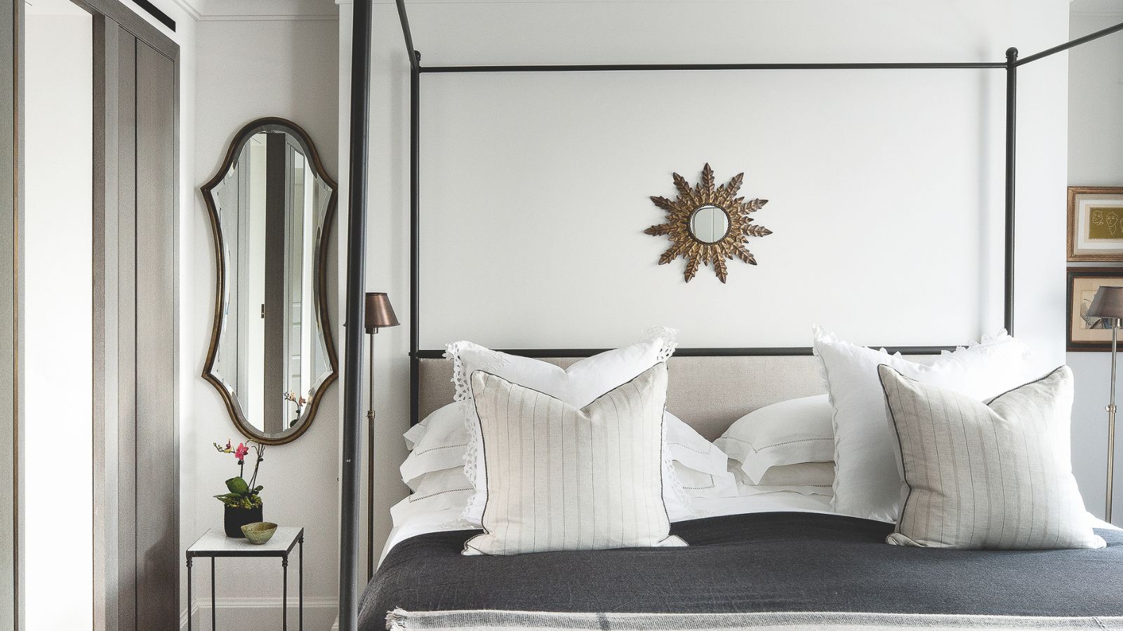 White bedroom with a black four-poster bed, grey bedding with white cushions, and a small circular mirror above the bed. To the left is an oval shaped mirror.