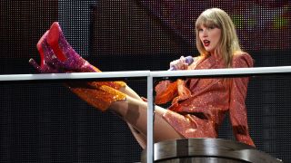 Taylor Swift in an orange and pink blazer putting her feet up on the railing while singing during the Eras Tour concert in Paris on May 12.