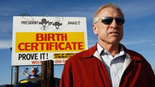 A man wearing sunglasses stands in front of a billboard. The billboard features a caricature of Barack Obama as a muslim and reads "President or Jihad? Birth Certificate: Prove it!"