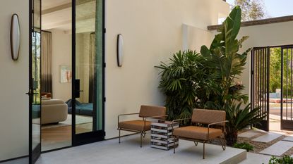 Image of a patio on the outside of a home that is painted a soft cream color. There are two orange outdoor chairs and an accent table between them. The door to inside is open.