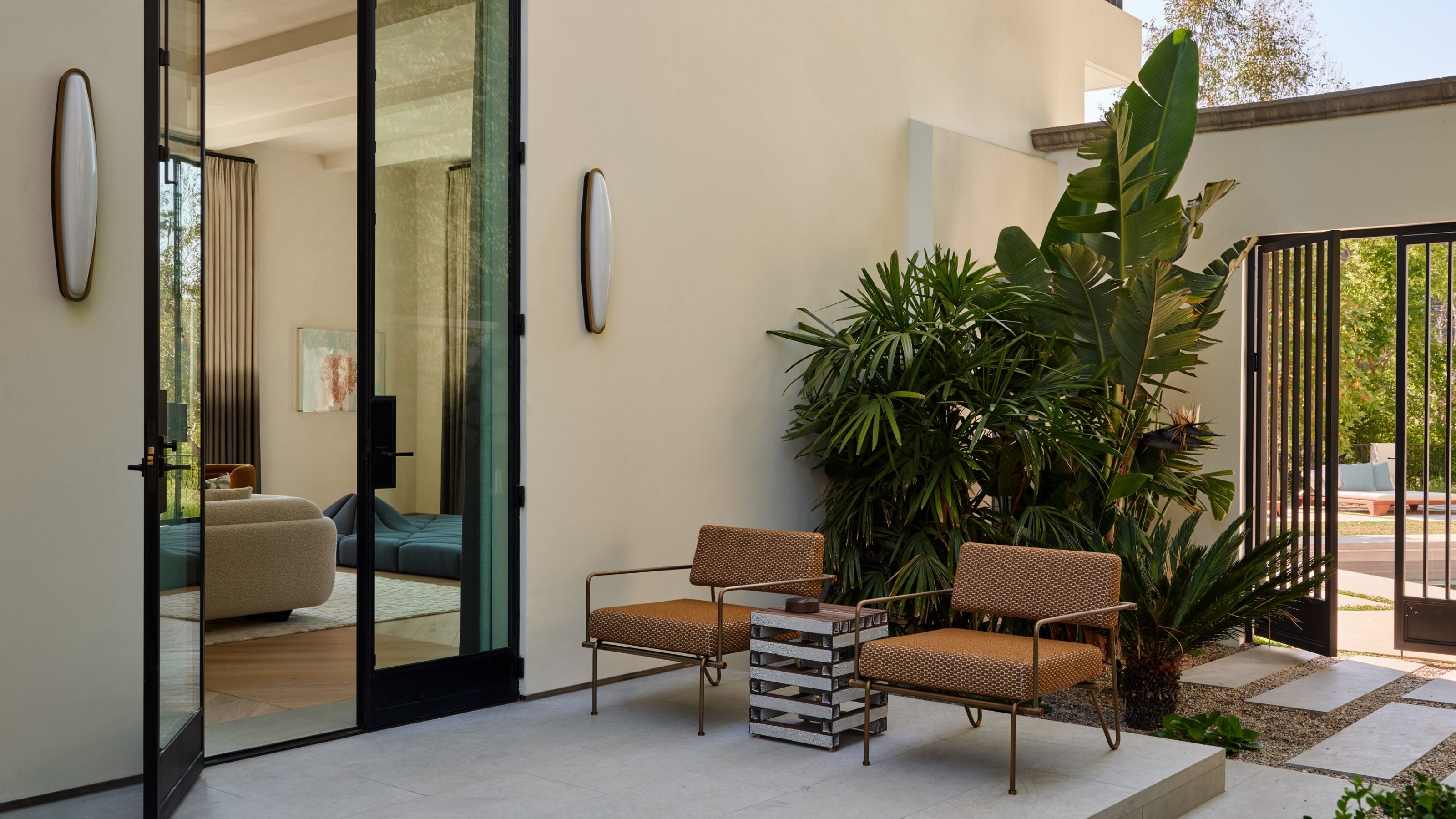 Image of a patio on the outside of a home that is painted a soft cream color. There are two orange outdoor chairs and an accent table between them. The door to inside is open.