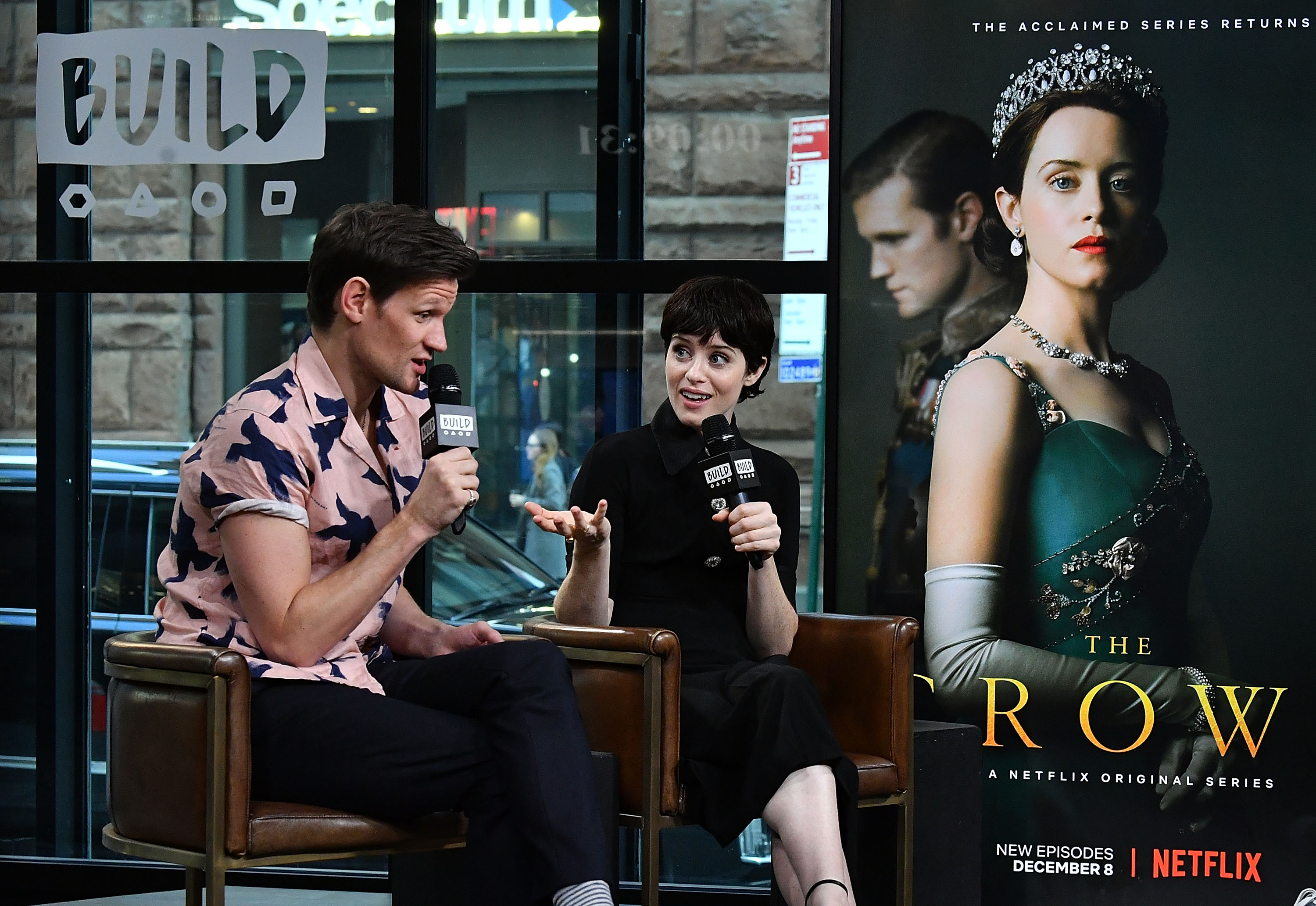 Matt Smith and Claire Foy sitting on chairs speaking into mics next to a poster for 'The Crown.'