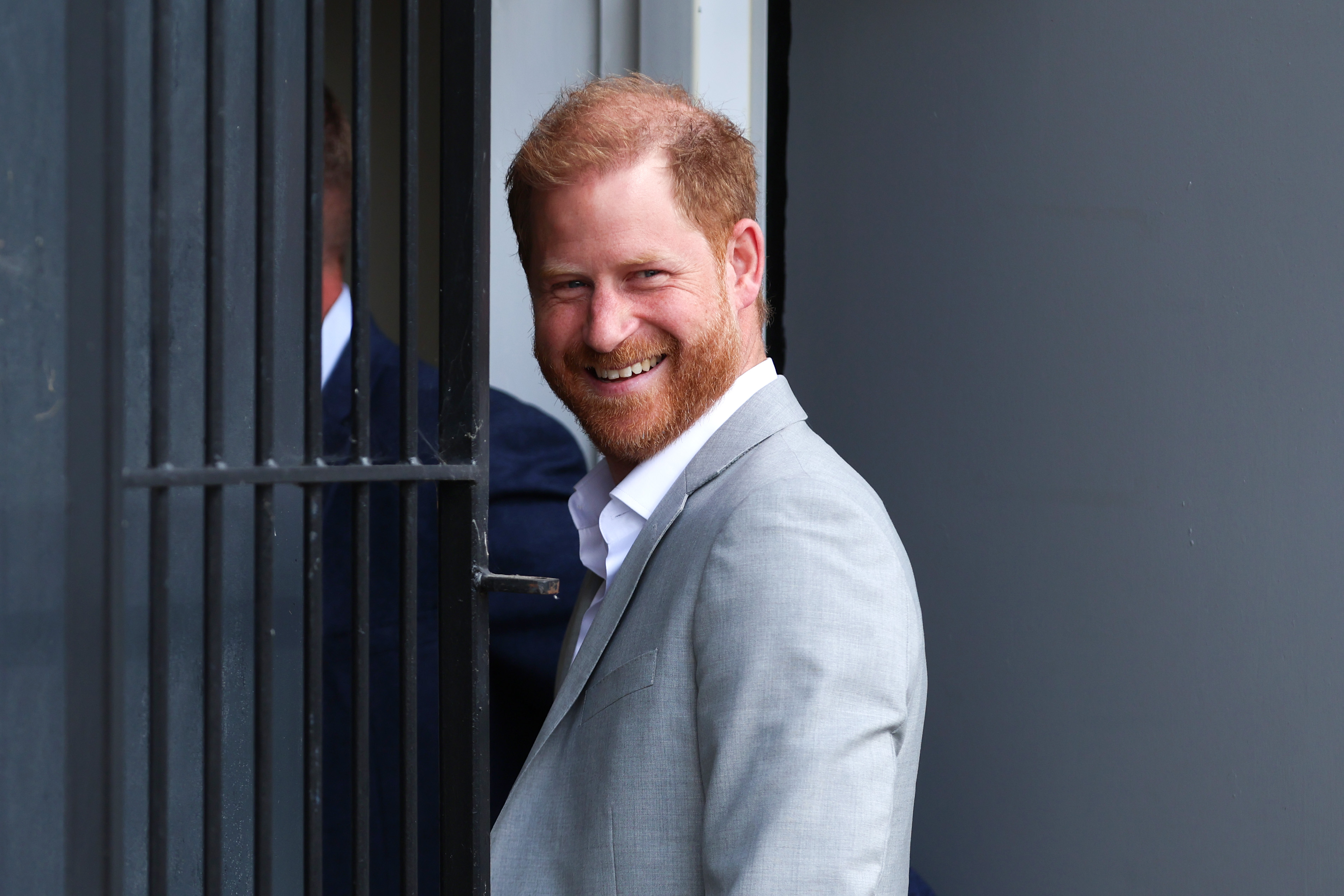 Prince Harry wears a gray suit jacket, a white shirt, and dark slacks and has red hair as he visits the UK in September 2025