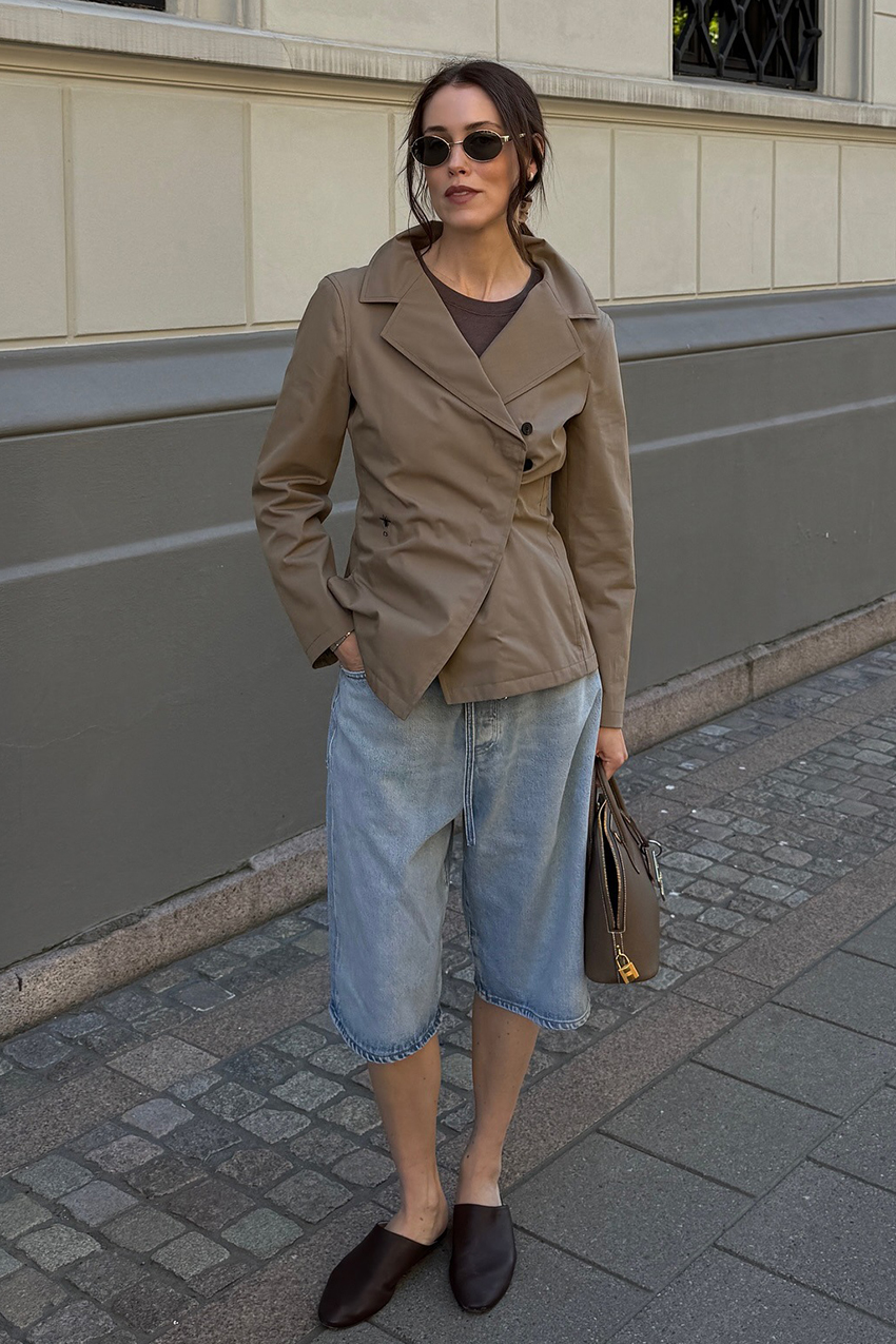 a woman wearing a cropped trench jacket, denim bermuda shorts, and black flats