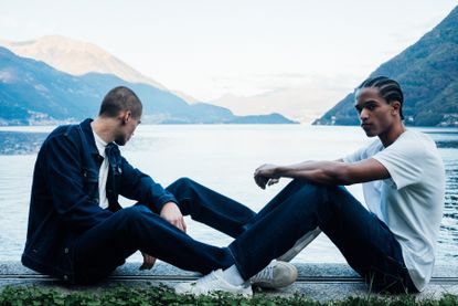Two men in Jacob Cohën jeans and T-shirts sat by lake with mountains in background