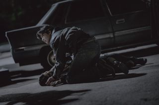 Park Seo-jun (as Jang Ho-jae) kneels over Han So-hee as (Yoon Chae-ok) as she lies on the ground in front of a car in Gyeongseong Creature S2.