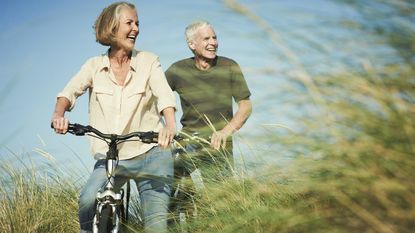 A happy older couple rides their bikes on a path through a green field.