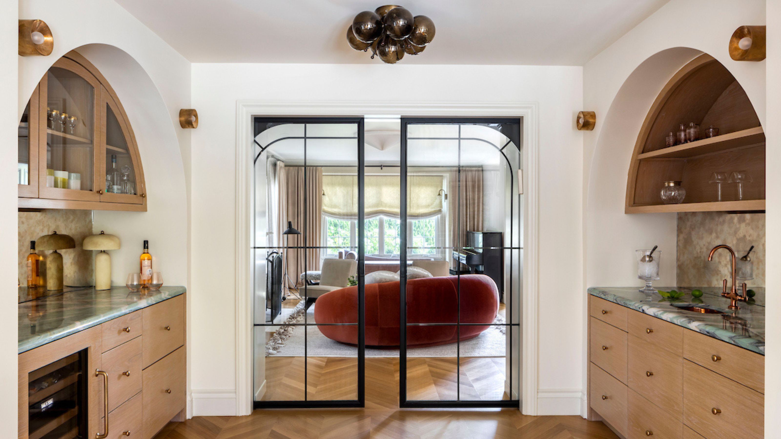 A view of one part of the kitchen that has doors leading onto the main living room area. The doors are panelled in a black frame and in the kitchen area, there is cabinetry and storage on either side with glass fronted cabinets as well as open shelving