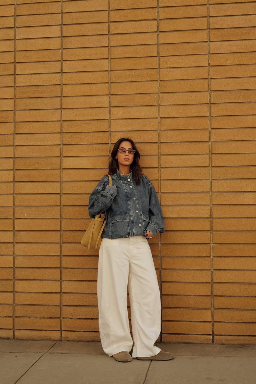 How to style white jeans for fall, shown in a photo of a woman standing outside against a yellow brick wall, wearing brown sunglasses, an oversized denim funnel neck jacket, a white t-shirt, white wide-leg jeans, suede Birkenstocks, and an oversized brown tote bag