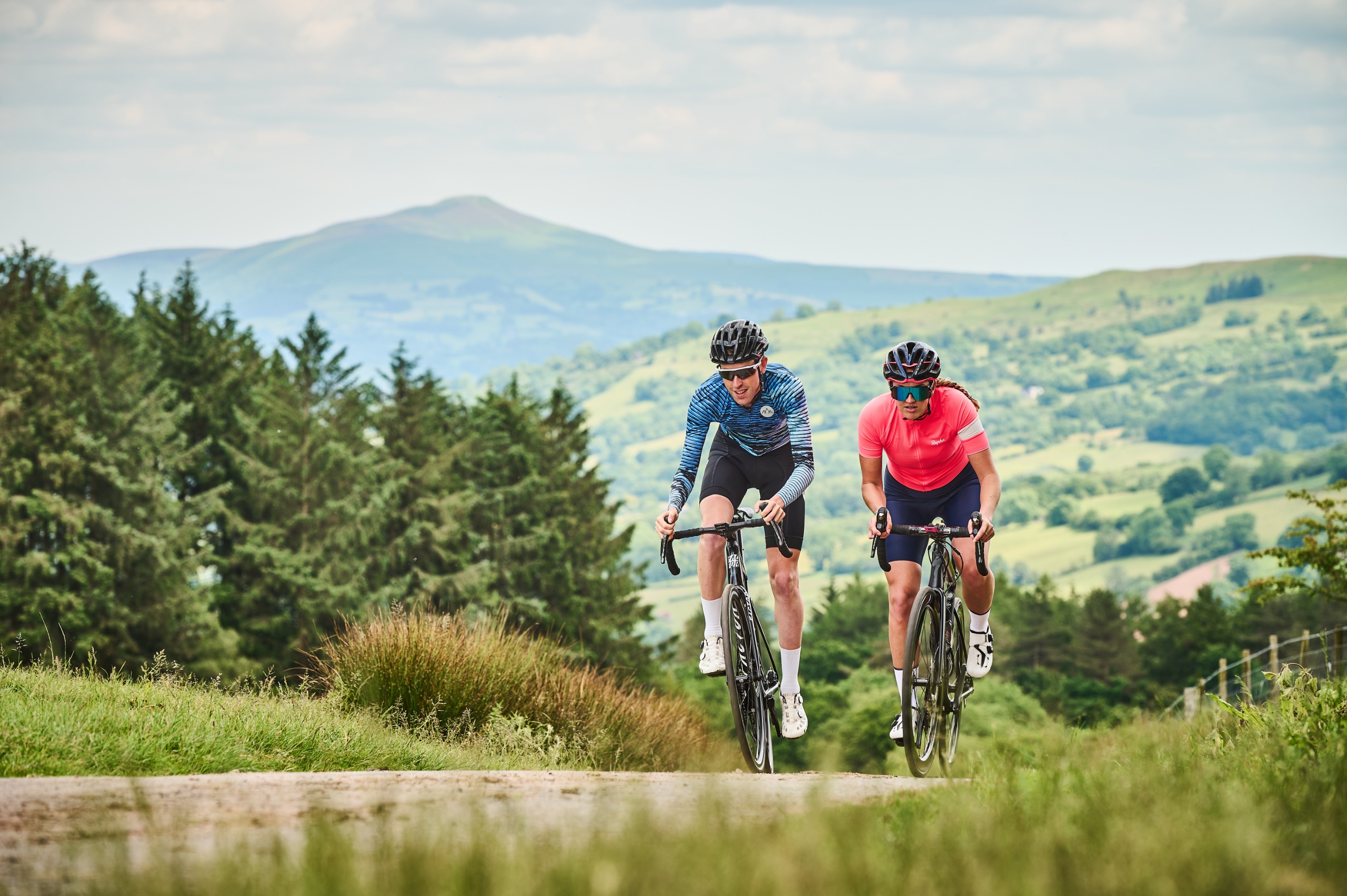 Two riders passing a forest, with hills in the background