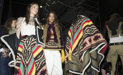 Models waiting for a fashion show to start, wearing colorful skirts and ponchos with bold patterns paired with white shirts and pants, from Dsquared2 A/W 2015 collection.