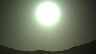 a bright, green-hued fireball of white hangs in the hazy sky above a low skyline of rolling hills.