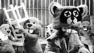 A black-and-white photo where a life-sized beaver holds a chair over its head, preparing to hit a man in a raccoon costume, in 'Hundreds of Beavers.'