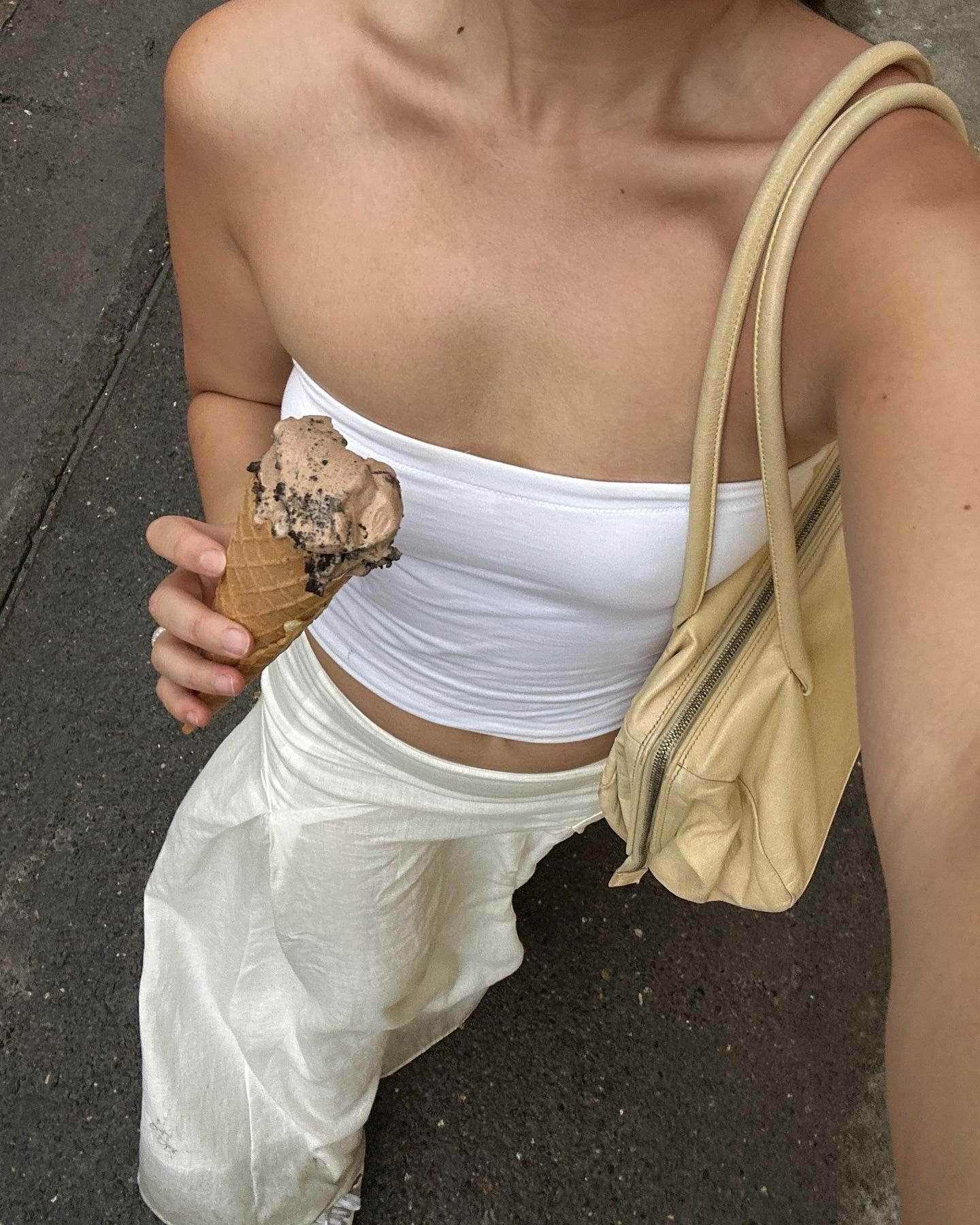 Influencer Jordan Risa takes a walking selfie while eating an ice cream cone wearing a butter-yellow shoulder bag, white tube top, and ivory linen skirt.