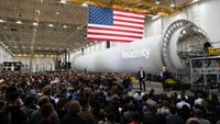 a large crowd sits facing to men in suits at one end of a rocket stage laid horizontally.