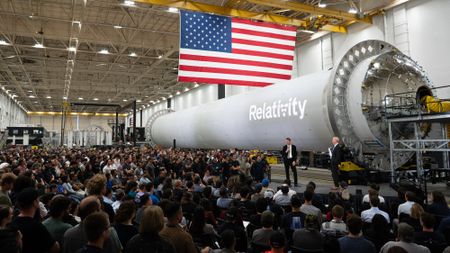 a large crowd sits facing to men in suits at one end of a rocket stage laid horizontally.