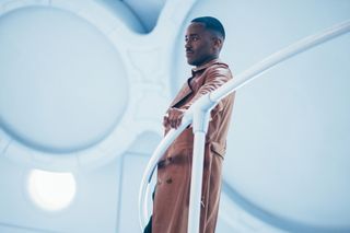 The Doctor (Ncuti Gatwa) stands on one of the gangways in the TARDIS, wearing a long brown leather coat and leaning on the handrail with his left arm