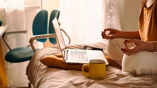 Woman doing yoga on the bed in sunshine with laptop and yellow mug of tea