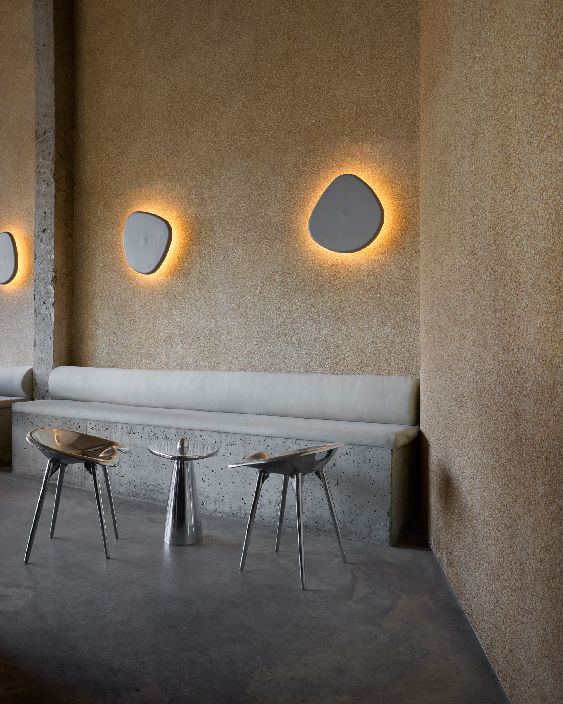 A brutalist building houses a sleek, almost utopian restaurant crafted from chocolatey slabs of stone, sheeny chrome, and exposed cement columns, with organically shaped, back-lit sconces resembling rocks.