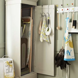 Neutral utility room, grey storage cupboard, storing vintage retro ironing board and cleaning accessories, hooks with various brushes, feather duster, dustpan and brush