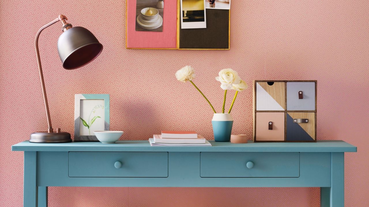 White and pink wall and rug, with corkboard and baby blue sideboard covered in objects in shades of blue, pink, yellow, and orange