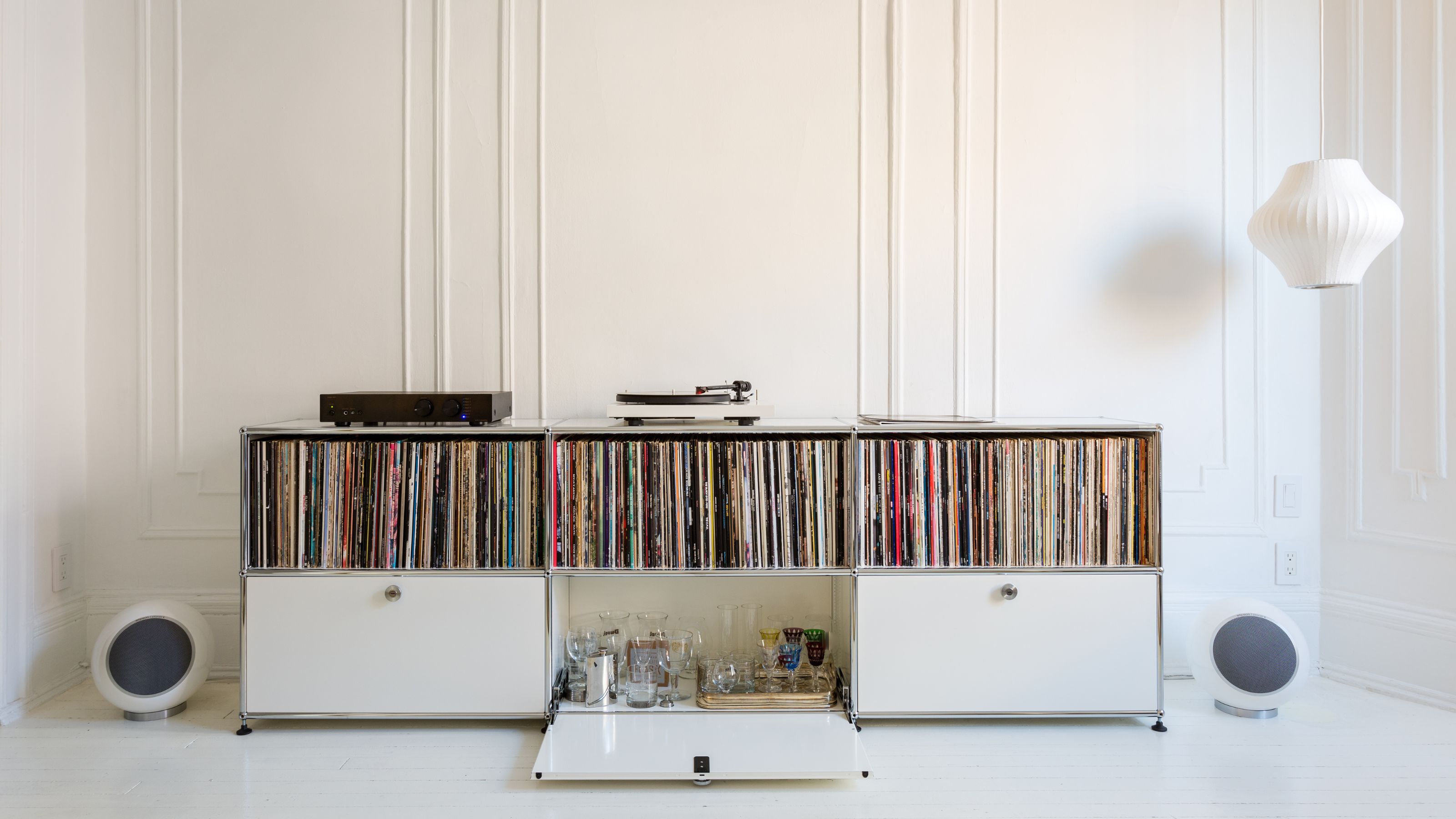 Image of an all-white room with a long and low record storage cabinet. It is full of records and there is an open drawer with cocktail glasses inside. 