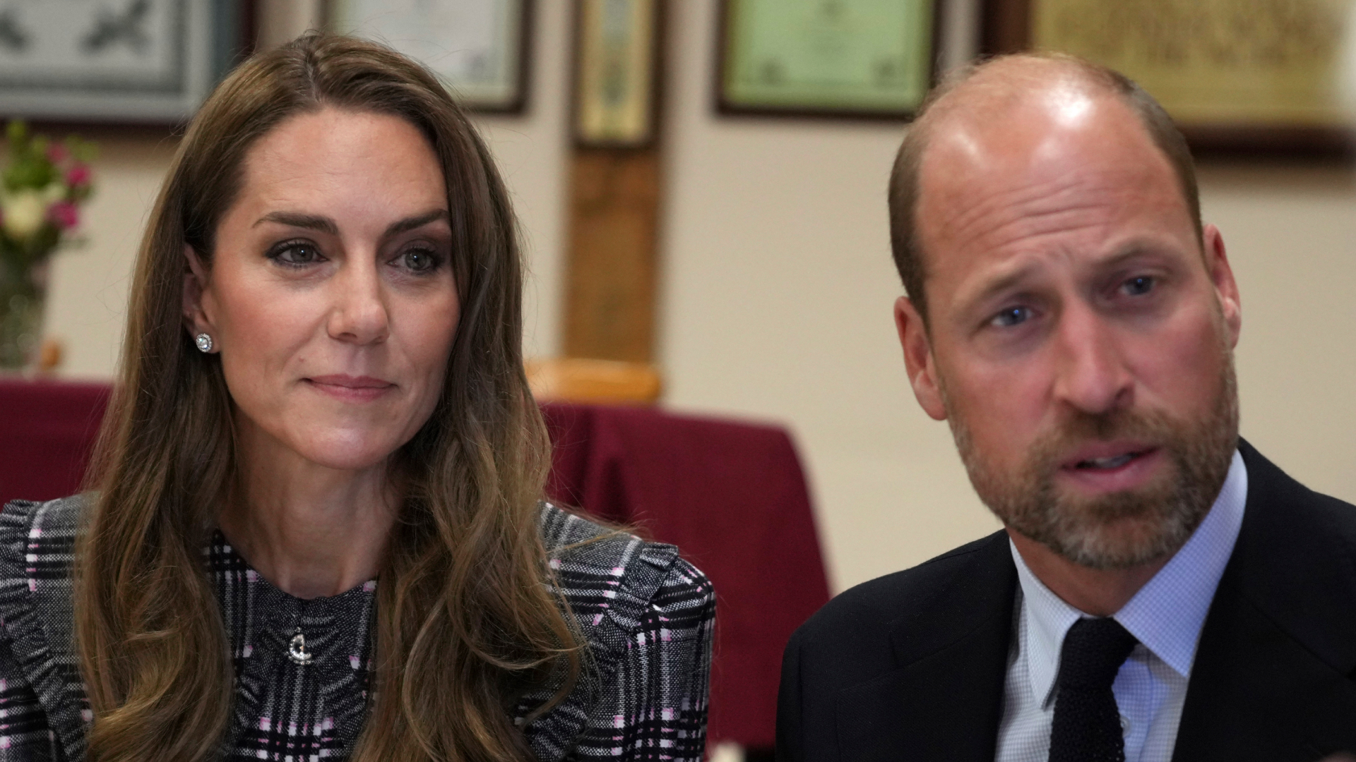 Kate Middleton wears a checked coat and has long wavy brunette hair while sitting next to Prince William