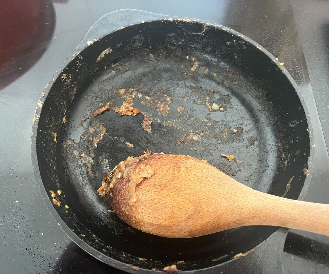 The stuck on remnants of food in a black cast iron skillet, with a wooden spoon.