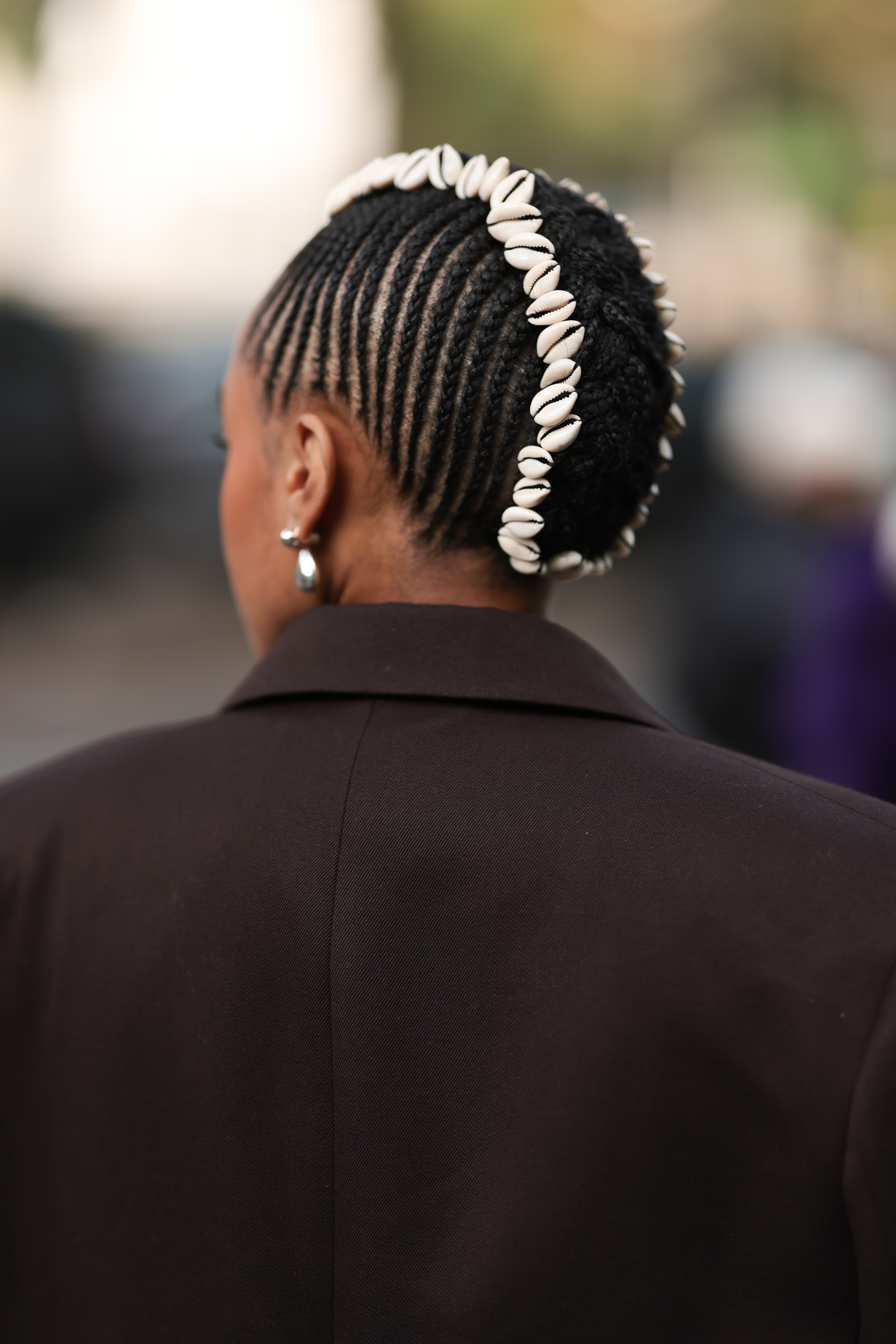 A woman wearing cowrie seashells in her fauxhawk cornrows hairstyle. She is also wearing a brown blazer.