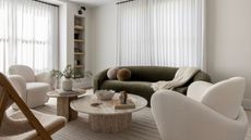 A white living room with cream accent chairs, a rounded olive green sofa and nested marble coffee tables.