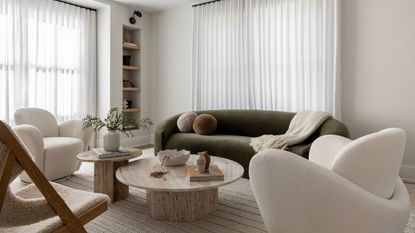 A white living room with cream accent chairs, a rounded olive green sofa and nested marble coffee tables.
