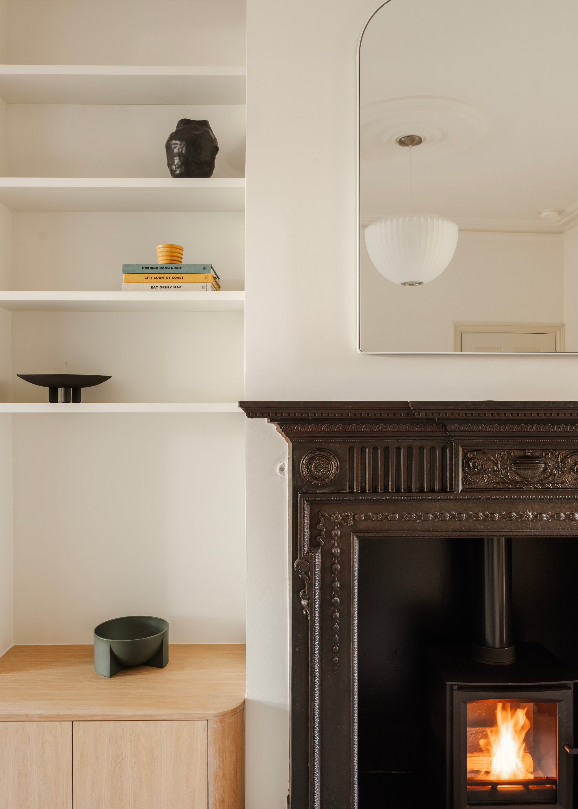 A living room with a beautifully carved fireplace, a pendant light, a curved mirror, open shelving with books, a pedestal bowl, a vase and a candle